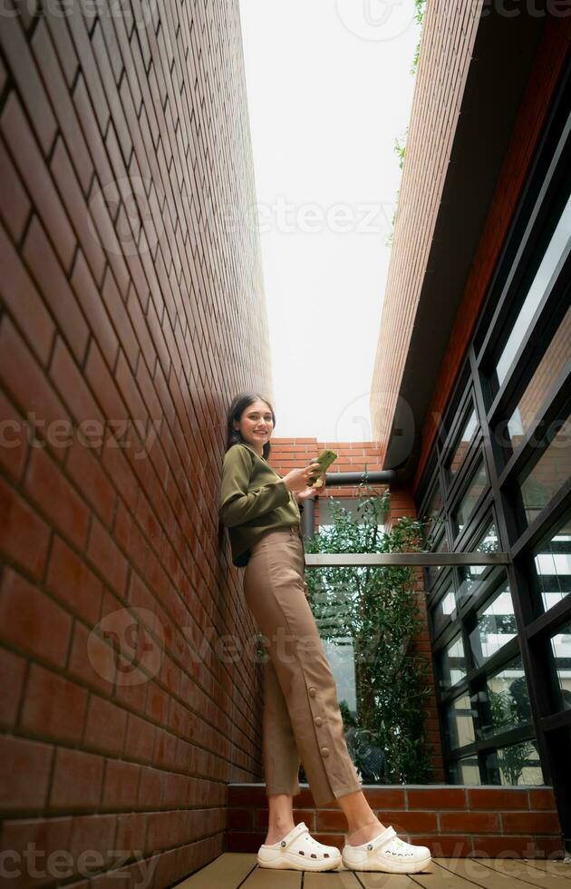 Portrait of a beautiful young woman drinking coffee and using mobile phone while leaning on a brick wall photo