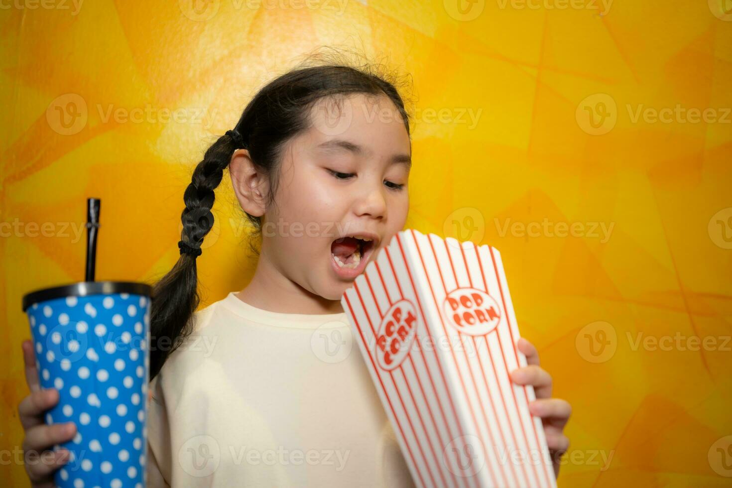 Asian child girl eating popcorn and drinking soda on yellow wall background, Cinema concept, photo