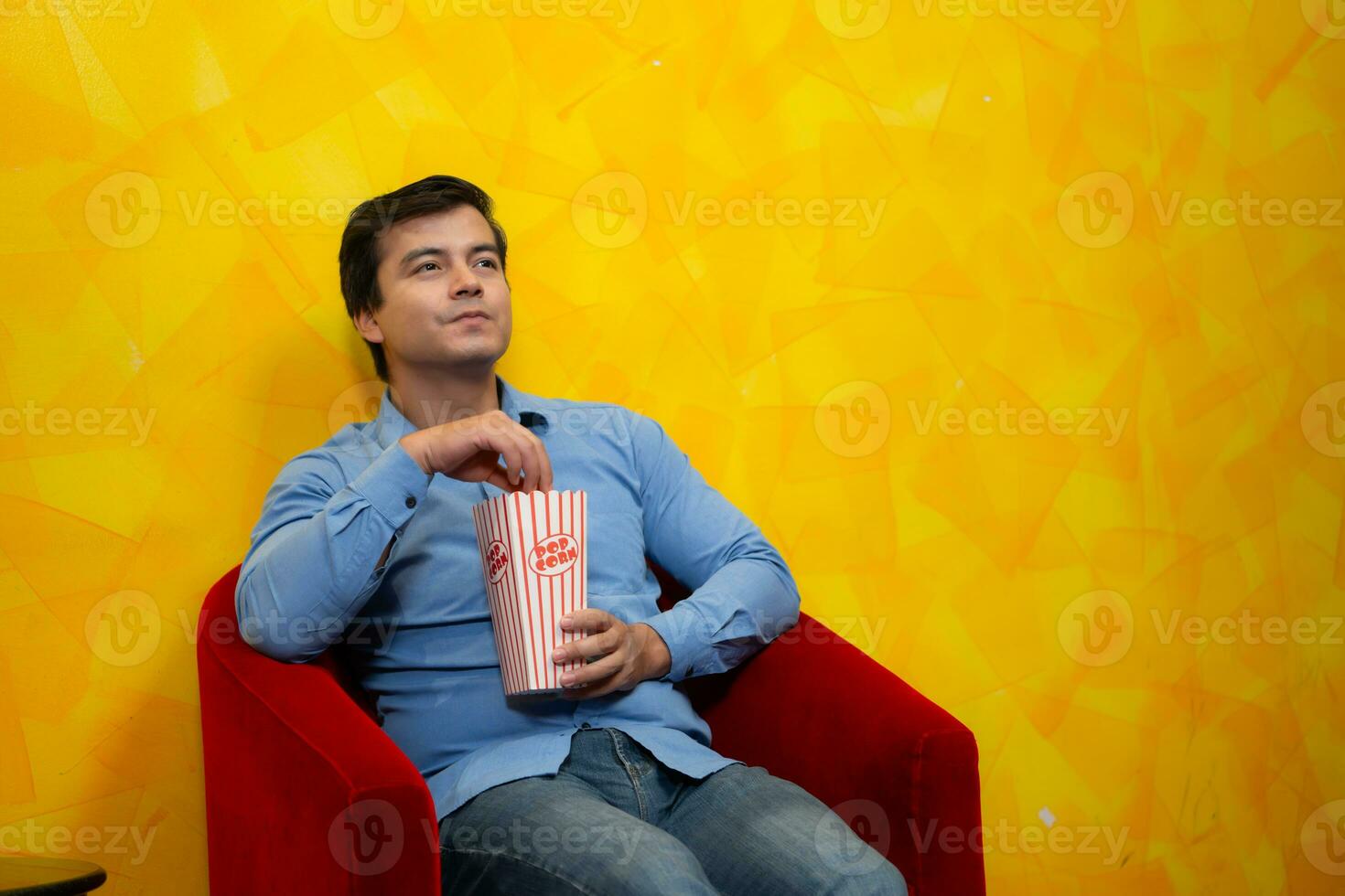 Young man eating popcorn while sitting in a red armchair against a yellow background waiting for watching a movie. photo