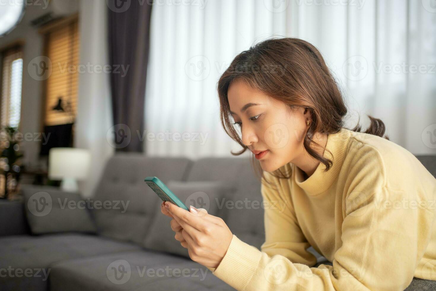 Happy young asian woman relax on sofa at home texting messaging on smartphone, smiling girl use smart phone chatting, browse wireless internet on gadget, shopping online from home photo