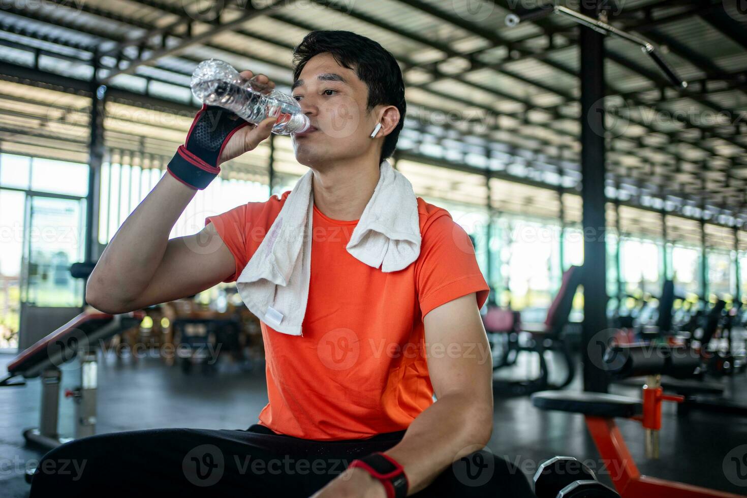 asiático carrocero hombre es sentado en banco y Bebiendo agua a relajarse después rutina de ejercicio en gimnasia. foto