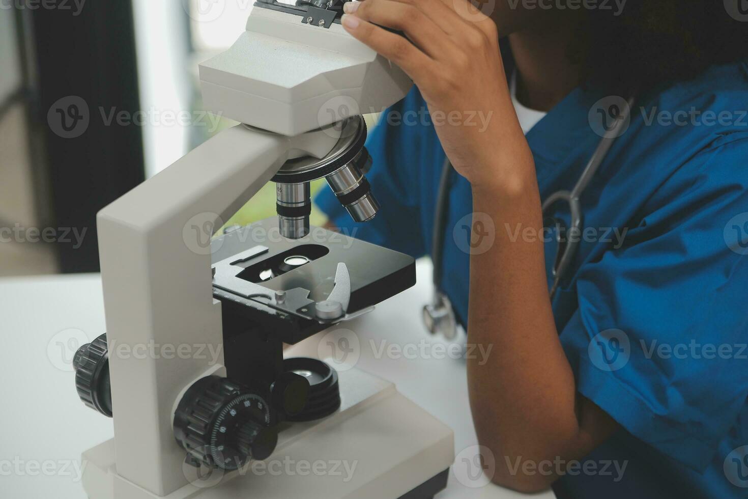 Medical worker in lab coat and sterile mask, doing a microscope analysis while her colleague are working behind photo