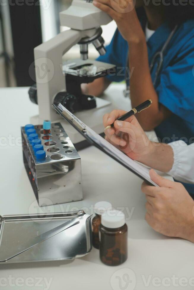 Medical worker in lab coat and sterile mask, doing a microscope analysis while her colleague are working behind photo