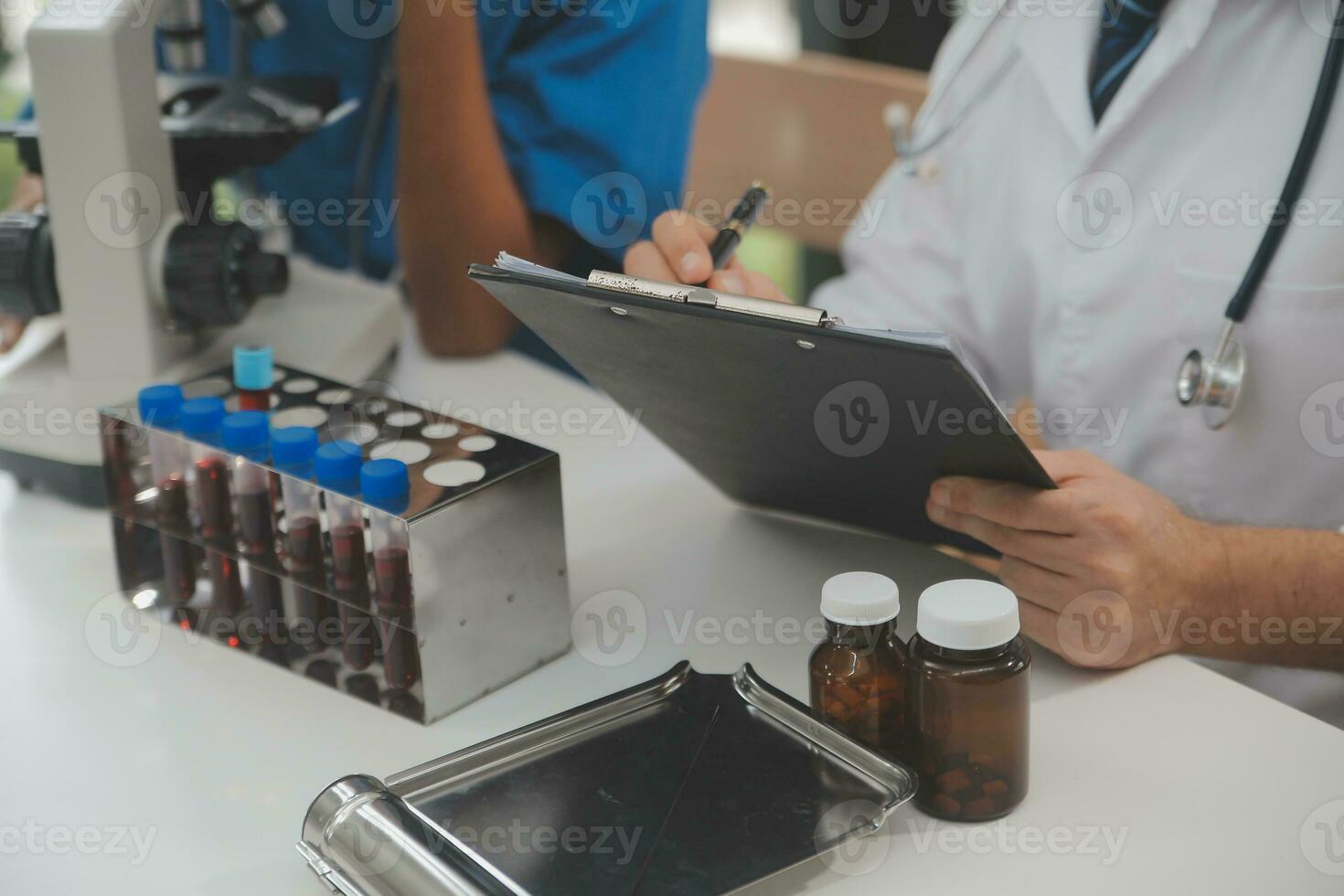 Medical worker in lab coat and sterile mask, doing a microscope analysis while her colleague are working behind photo
