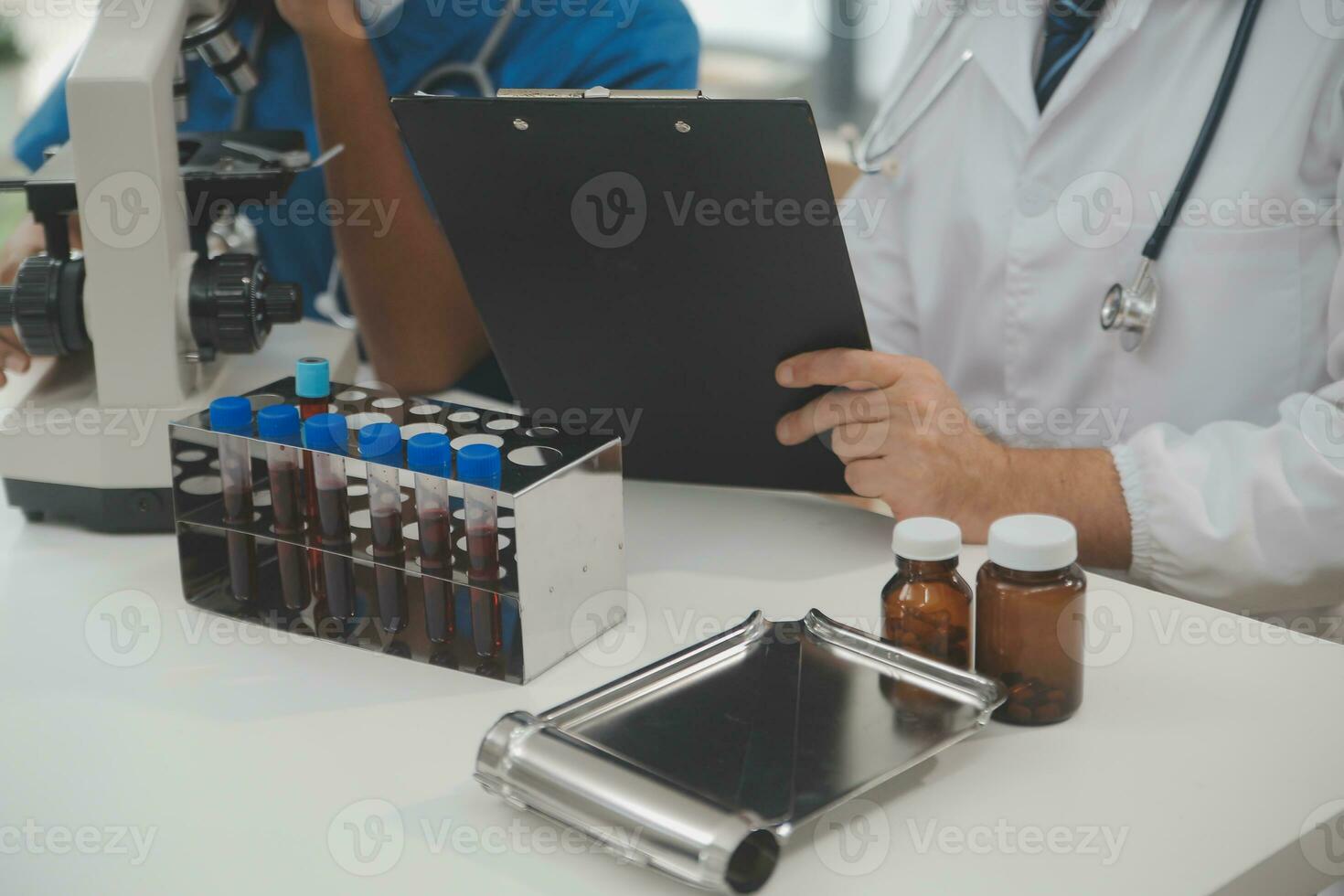 Medical worker in lab coat and sterile mask, doing a microscope analysis while her colleague are working behind photo