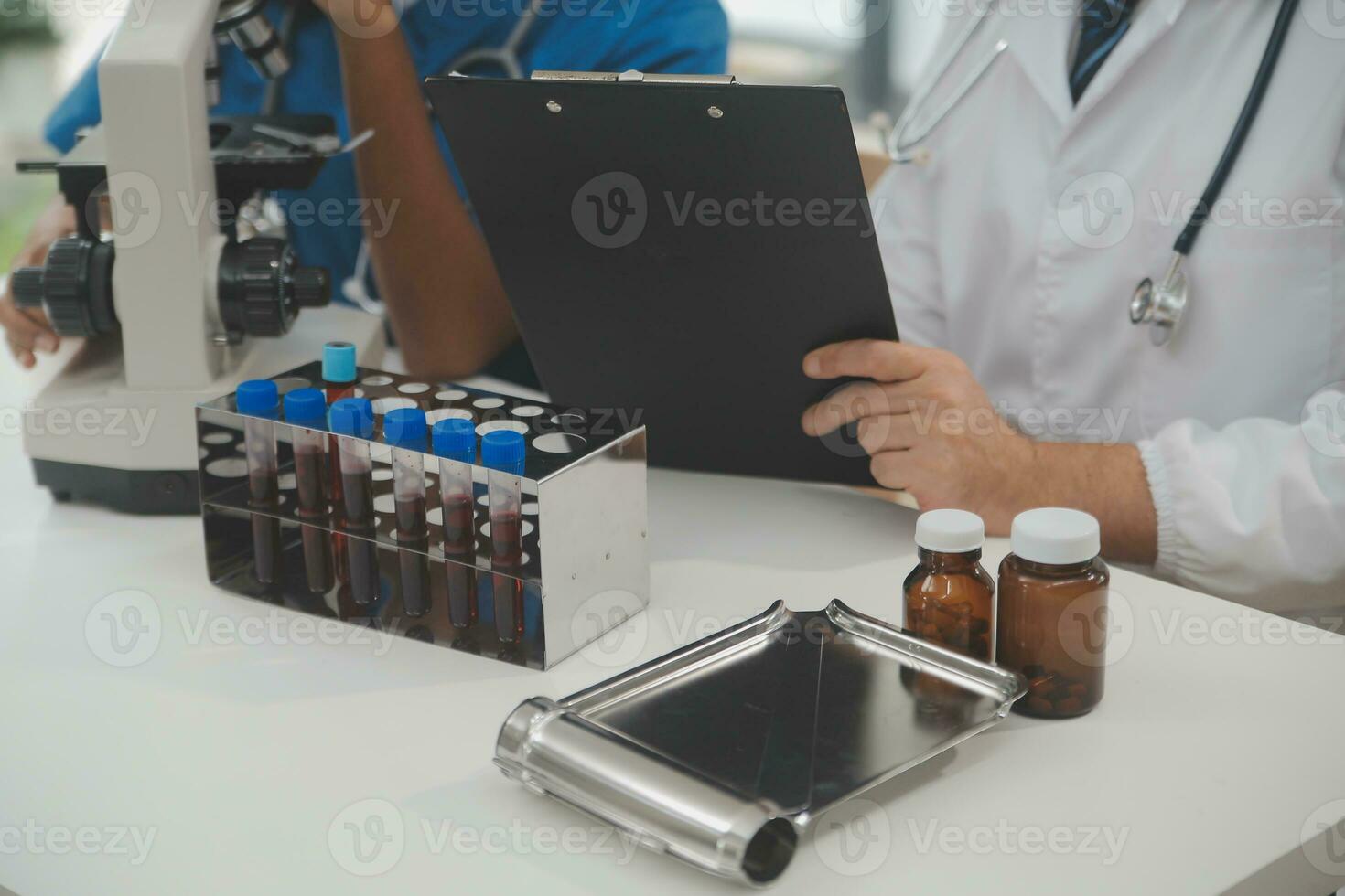 Medical worker in lab coat and sterile mask, doing a microscope analysis while her colleague are working behind photo