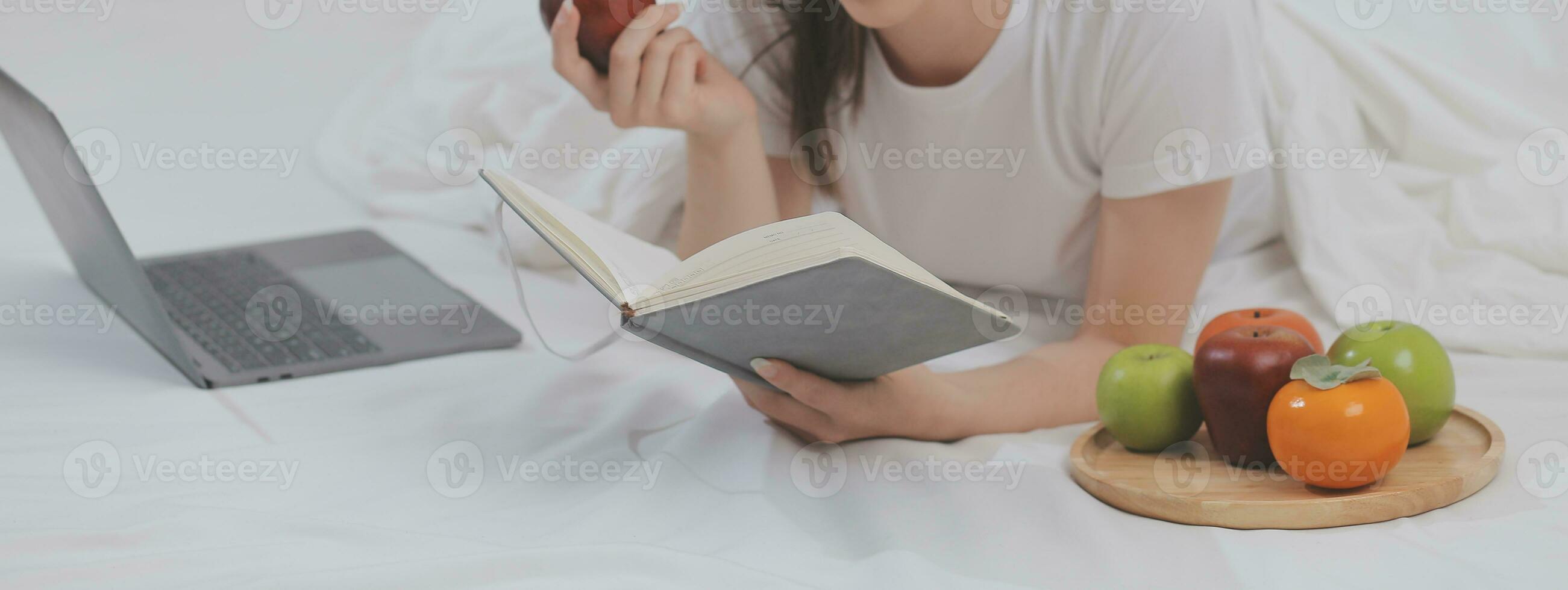 Efficient Young Millennial Girl Sitting on a Bed in the Morning, Uses Laptop Computer and Eats Croissants and Drinks Coffee for Breakfast photo