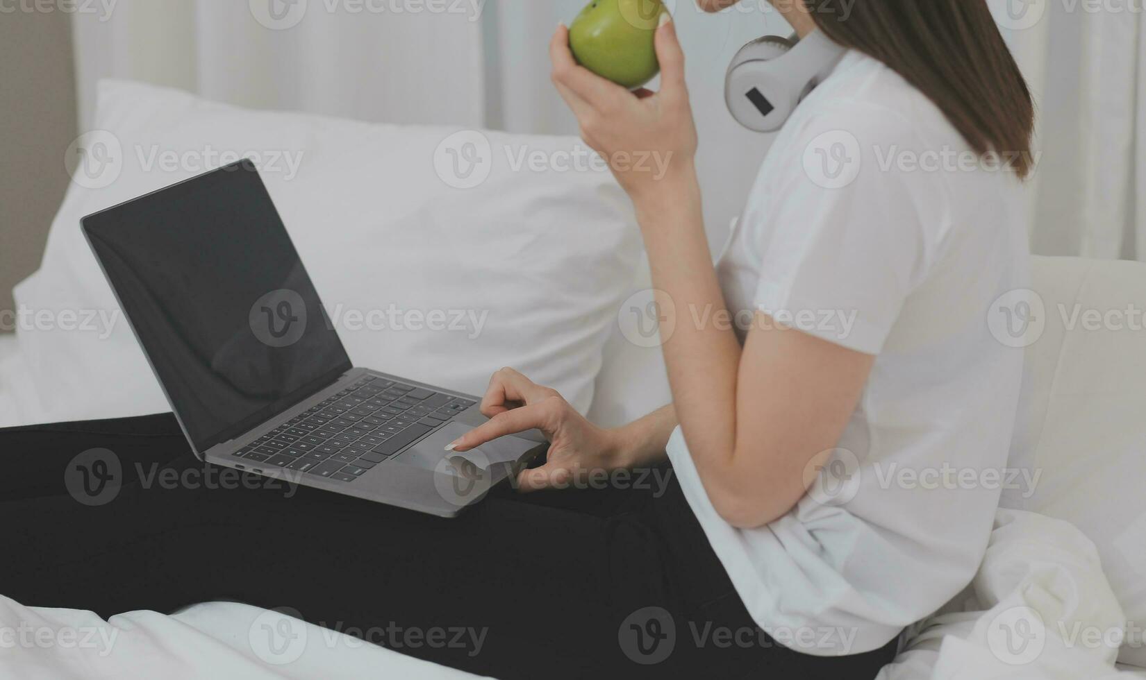 Efficient Young Millennial Girl Sitting on a Bed in the Morning, Uses Laptop Computer and Eats Croissants and Drinks Coffee for Breakfast photo