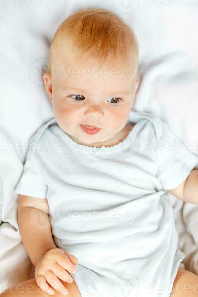Cute little newborn girl with smiling face looking at camera on white background. Infant baby resting playing lying down on crib bed at home. Motherhood happy child concept. photo