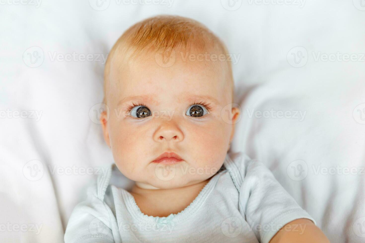 Cute little newborn girl with funny face looking at camera on white background. Infant baby resting playing lying down on crib bed at home. Motherhood happy child concept. photo