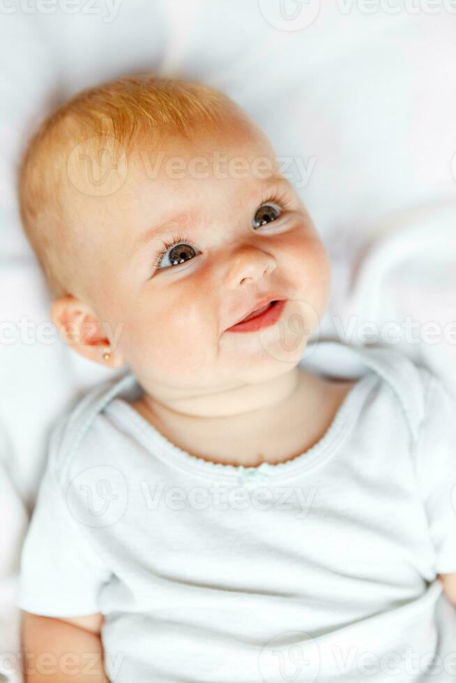 Cute little newborn girl with smiling face looking at camera on white background. Infant baby resting playing lying down on crib bed at home. Motherhood happy child concept. photo