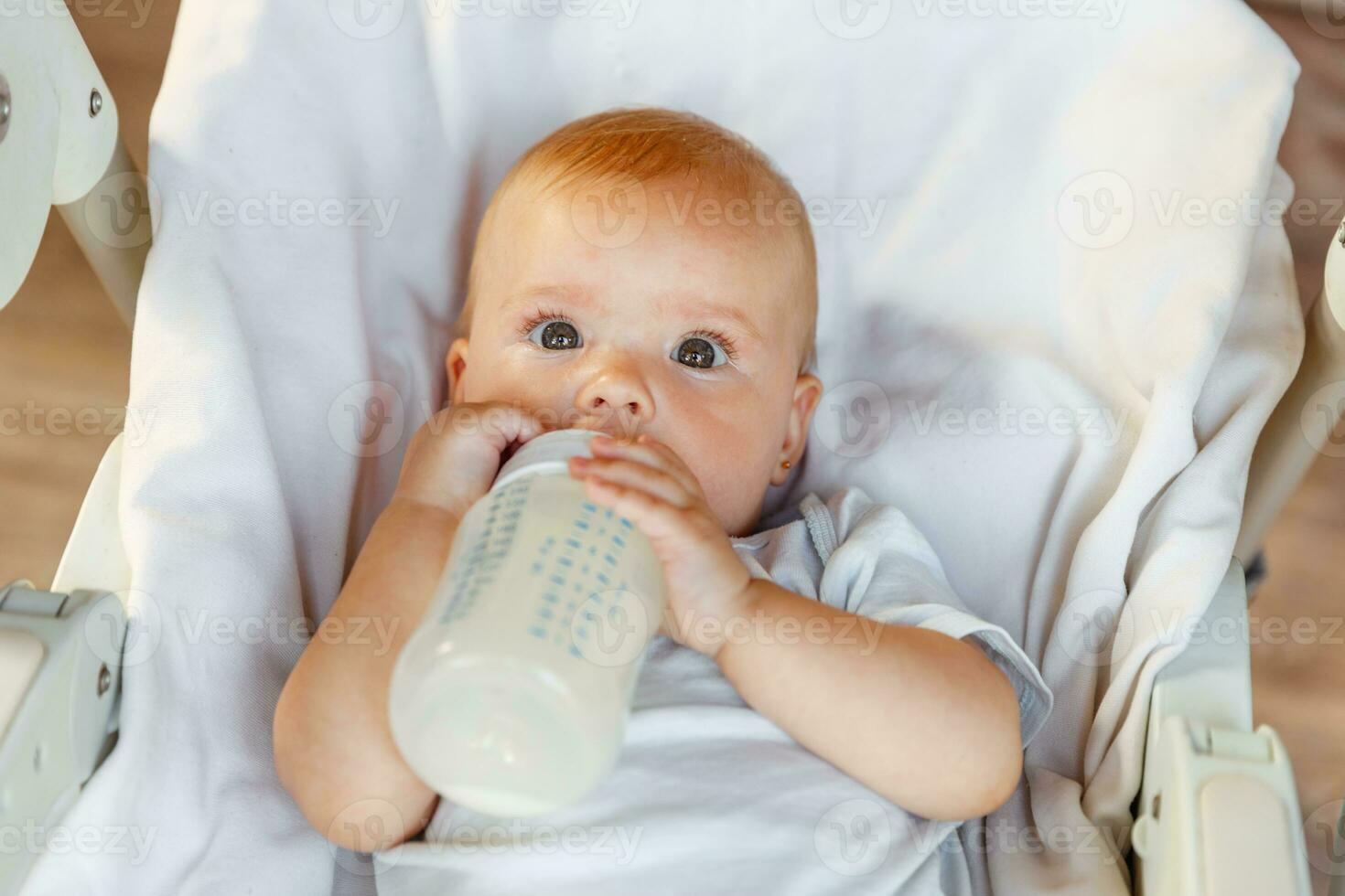 linda pequeño recién nacido niña Bebiendo Leche desde botella y mirando a cámara en blanco antecedentes. infantil bebé succión comiendo Leche nutrición acostado abajo en alimentación silla a hogar. maternidad contento niño foto