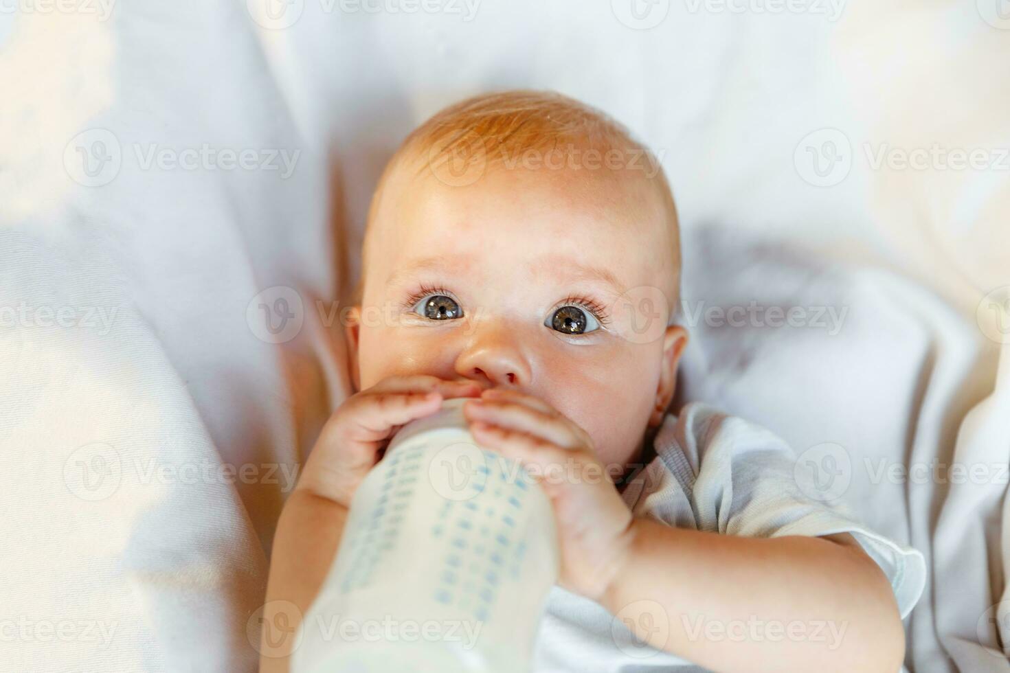 Cute little newborn girl drinking milk from bottle and looking at camera on white background. Infant baby sucking eating milk nutrition lying down on crib bed at home. Motherhood happy child concept. photo