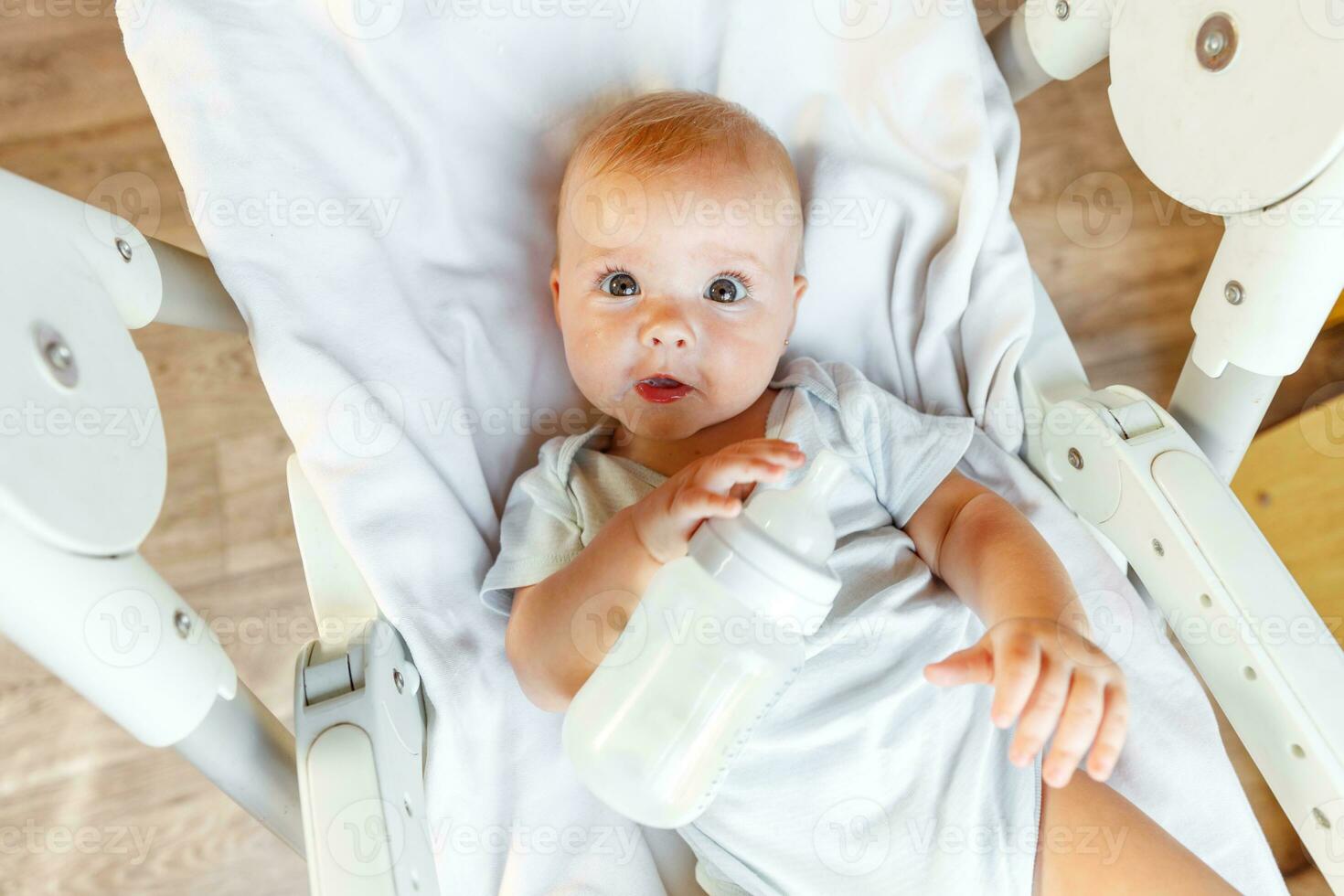 Cute little newborn girl drinking milk from bottle and looking at camera on white background. Infant baby sucking eating milk nutrition lying down on feeding chair at home. Motherhood happy child photo