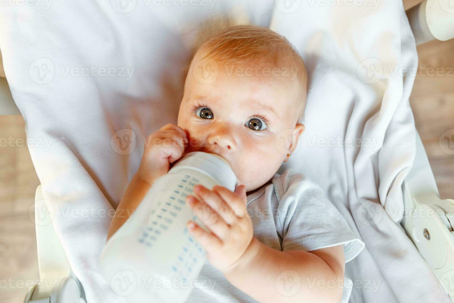 linda pequeño recién nacido niña Bebiendo Leche desde botella y mirando a cámara en blanco antecedentes. infantil bebé succión comiendo Leche nutrición acostado abajo en cuna cama a hogar. maternidad contento niño concepto. foto