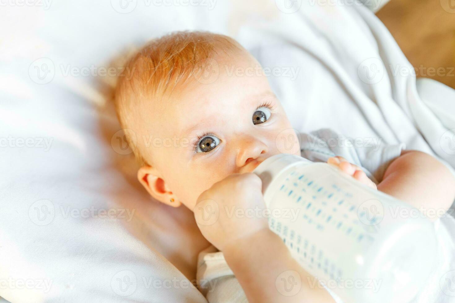 Cute little newborn girl drinking milk from bottle and looking at camera on white background. Infant baby sucking eating milk nutrition lying down on crib bed at home. Motherhood happy child concept. photo