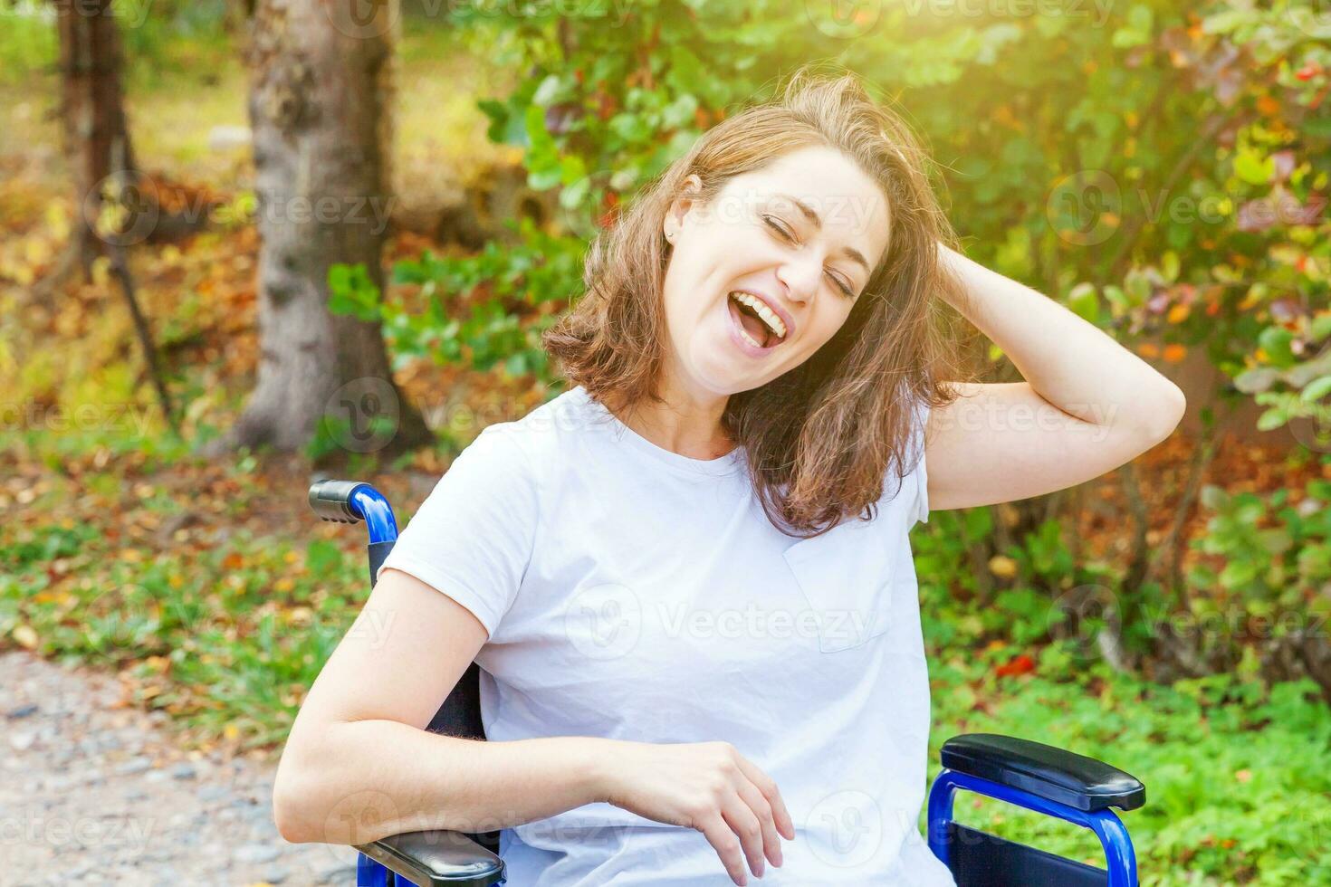 Young happy handicap woman in wheelchair on road in hospital park enjoying freedom. Paralyzed girl in invalid chair for disabled people outdoor in nature. Rehabilitation concept. photo