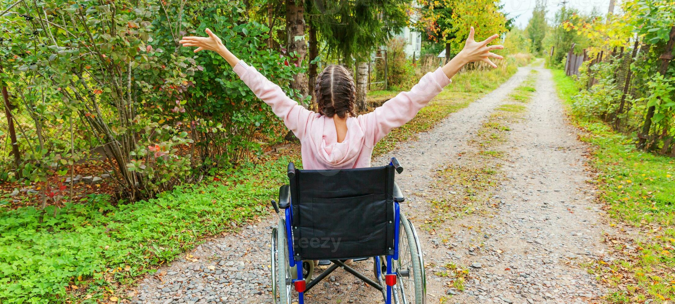 Young happy handicap woman in wheelchair on road in hospital park enjoying freedom. Paralyzed girl in invalid chair for disabled people outdoor in nature. Rehabilitation concept. photo