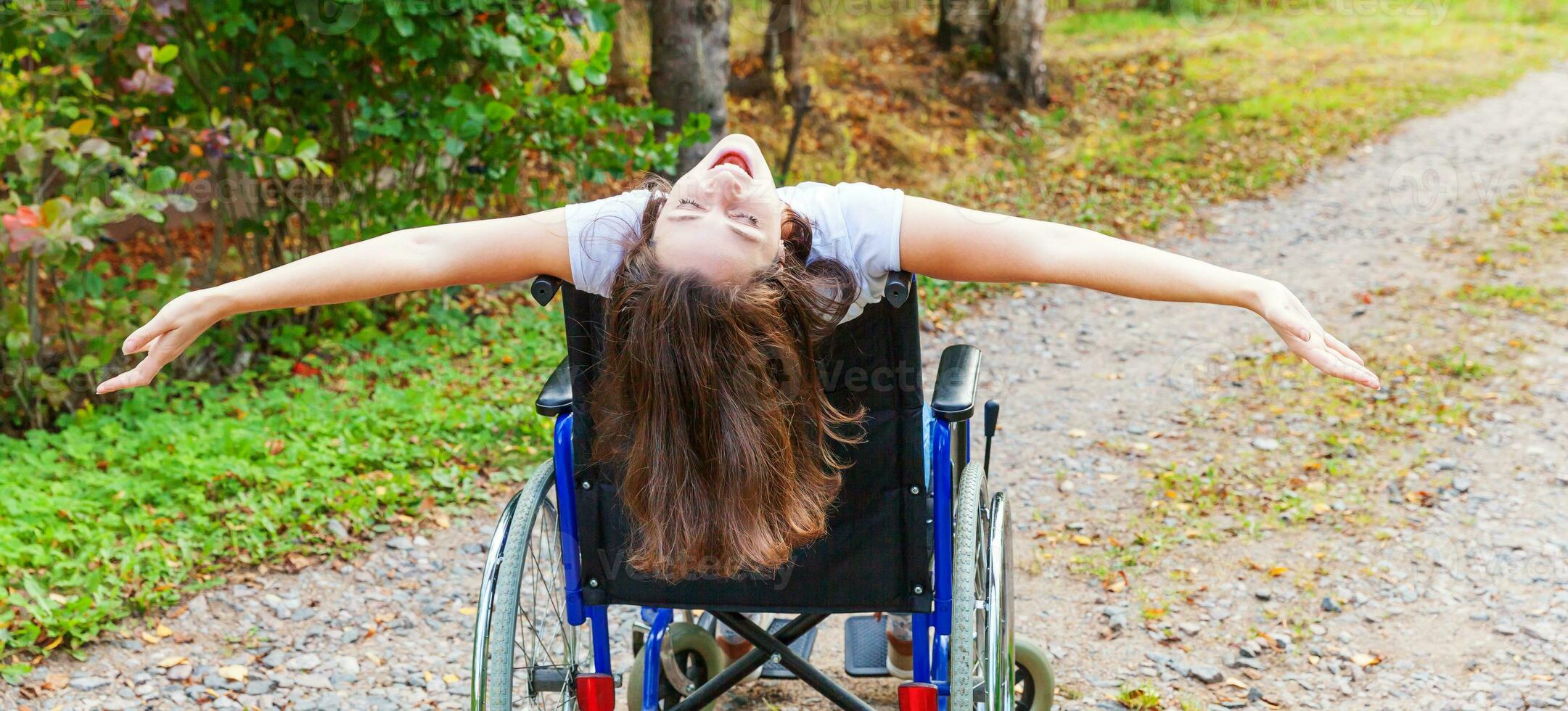 Young happy handicap woman in wheelchair on road in hospital park enjoying freedom. Paralyzed girl in invalid chair for disabled people outdoor in nature. Rehabilitation concept. photo