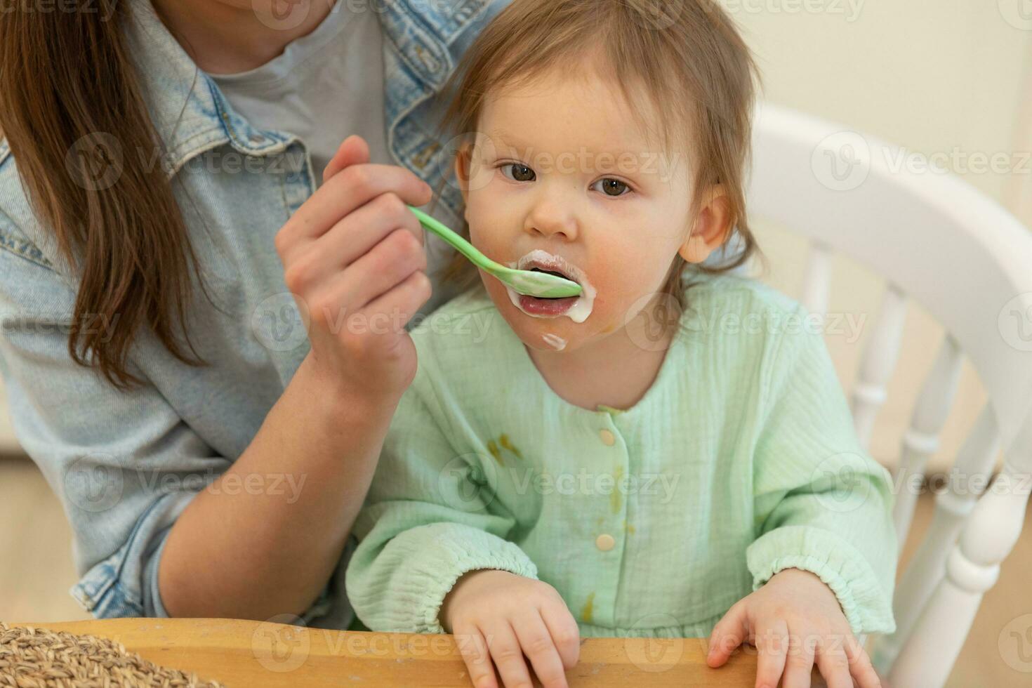 Happy family at home. Mother feeding her baby girl from spoon in kitchen. Little toddler child with messy funny face eats healthy food at home. Young woman mom giving food to kid daughter. photo