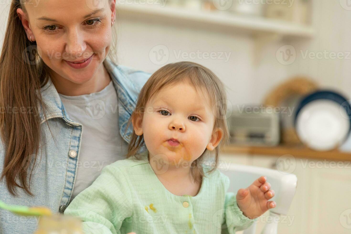 Happy family at home. Mother feeding her baby girl from spoon in kitchen. Little toddler child with messy funny face eats healthy food at home. Young woman mom giving food to kid daughter. photo