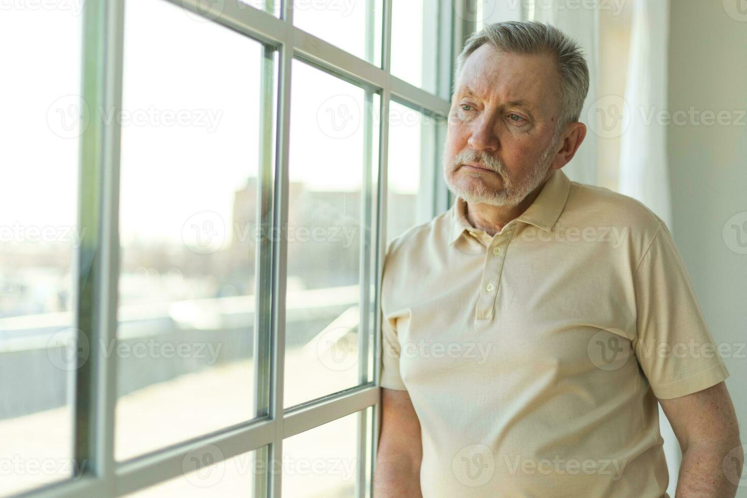 Sad tired lonely disappointed older senior man at home alone. Unhappy grandfather looking sadly outside window experiencing grief relative death bad news. Stressed elder man suffering from loneliness. photo