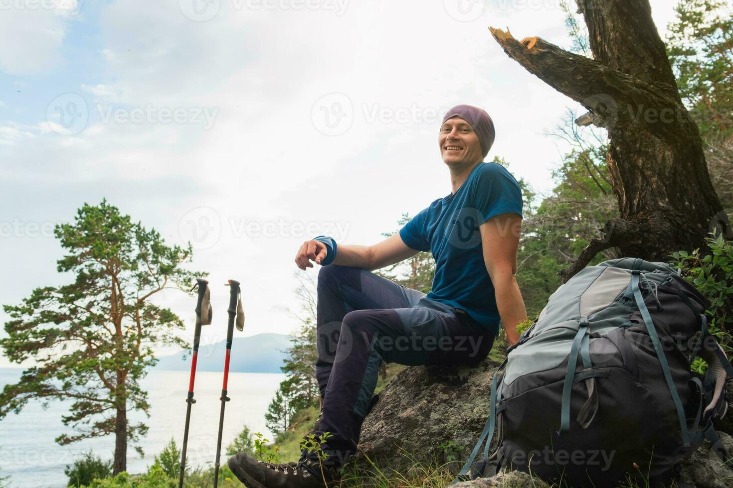 Hiking climbing cliff adventure. Backpacker man looking at beautiful view. Hiker with backpack sits on big rock above green forest and lake. Young happy hiker man smiling enjoy hike trekking tourism. photo
