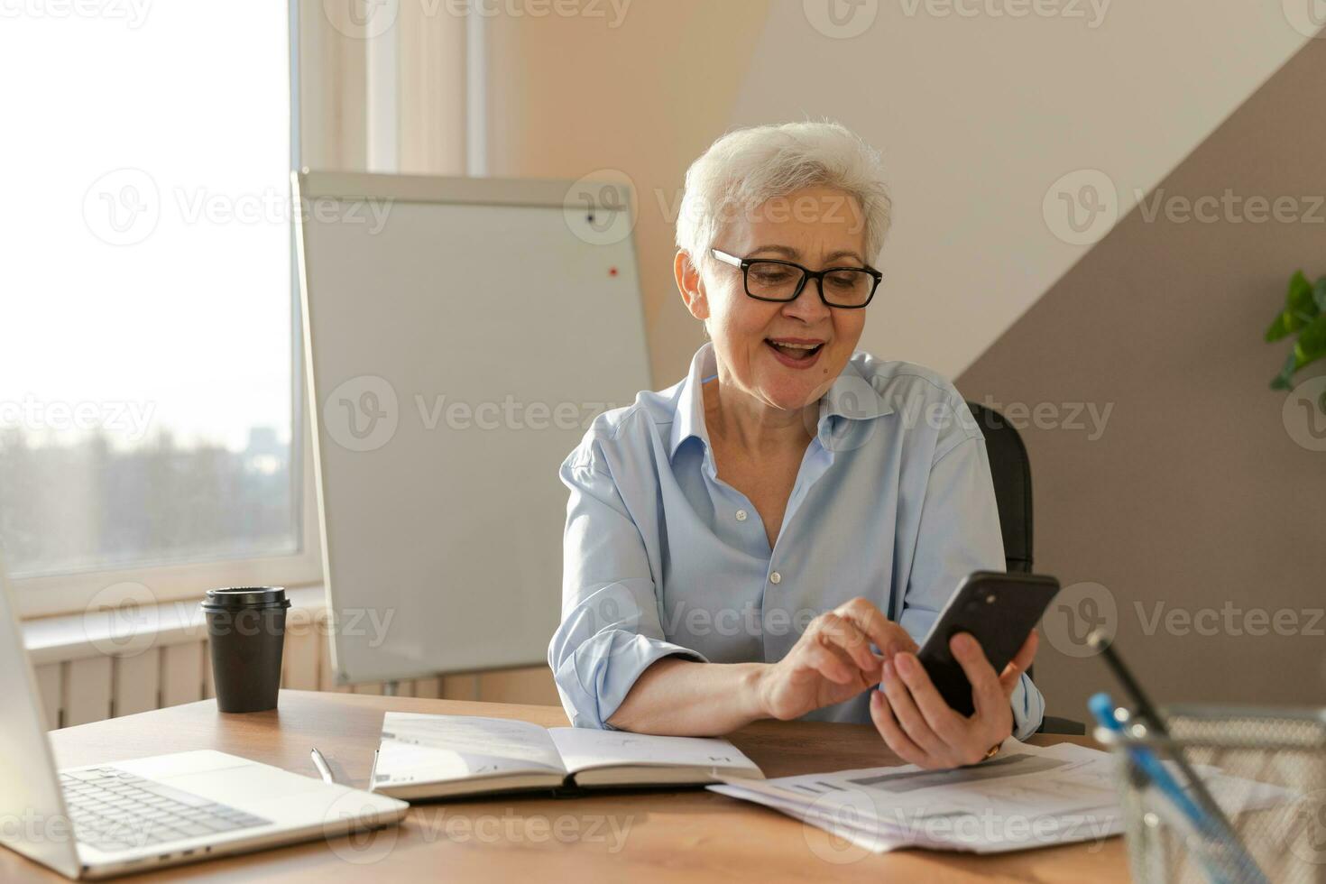 confidente elegante europeo medio Envejecido mayor mujer utilizando teléfono inteligente a lugar de trabajo. elegante más viejo maduro 60s gris peludo dama mujer de negocios con célula teléfono en oficina. jefe líder utilizando Internet aplicaciones foto