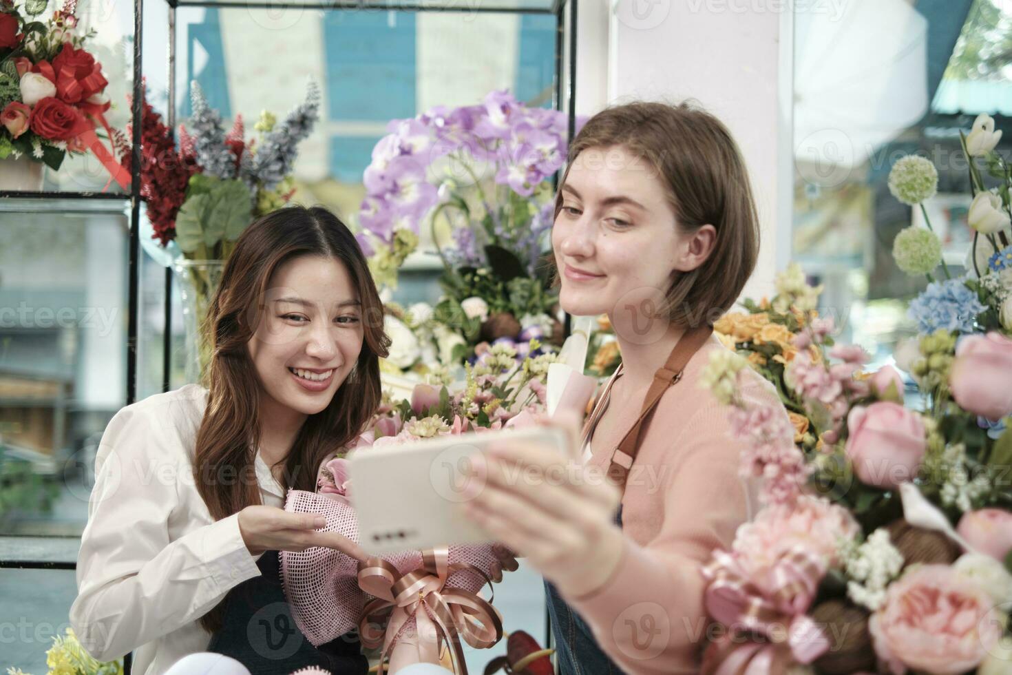 E-commerce business partner, two young female florist workers show floral arrangement, online live streaming, and selfies with smartphone application in bright flower shop, beautiful blossoms store. photo