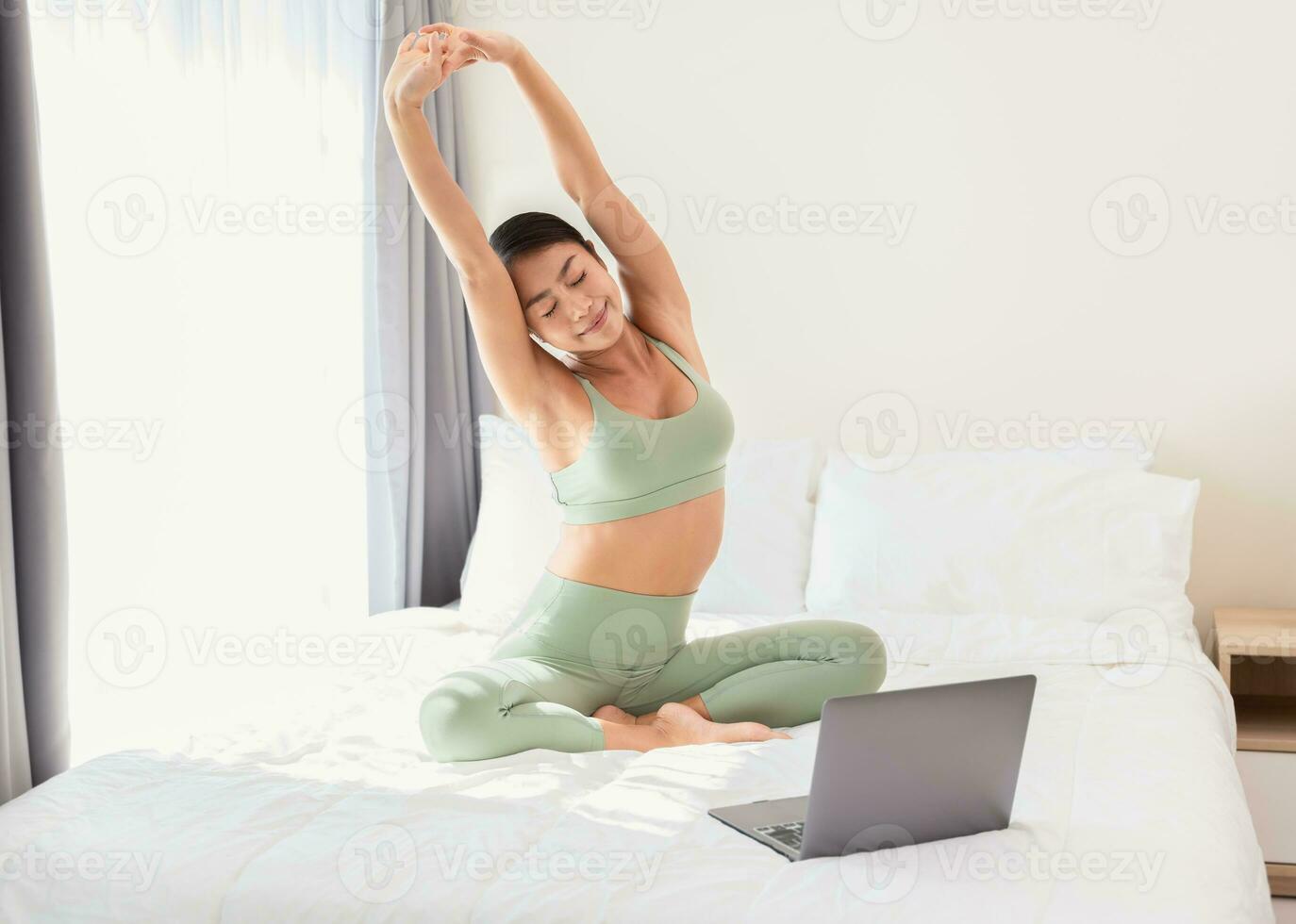 Healthy woman stretching on bed with arms raised up, preparing the body for yoga training. Online yoga training in morning at home. photo