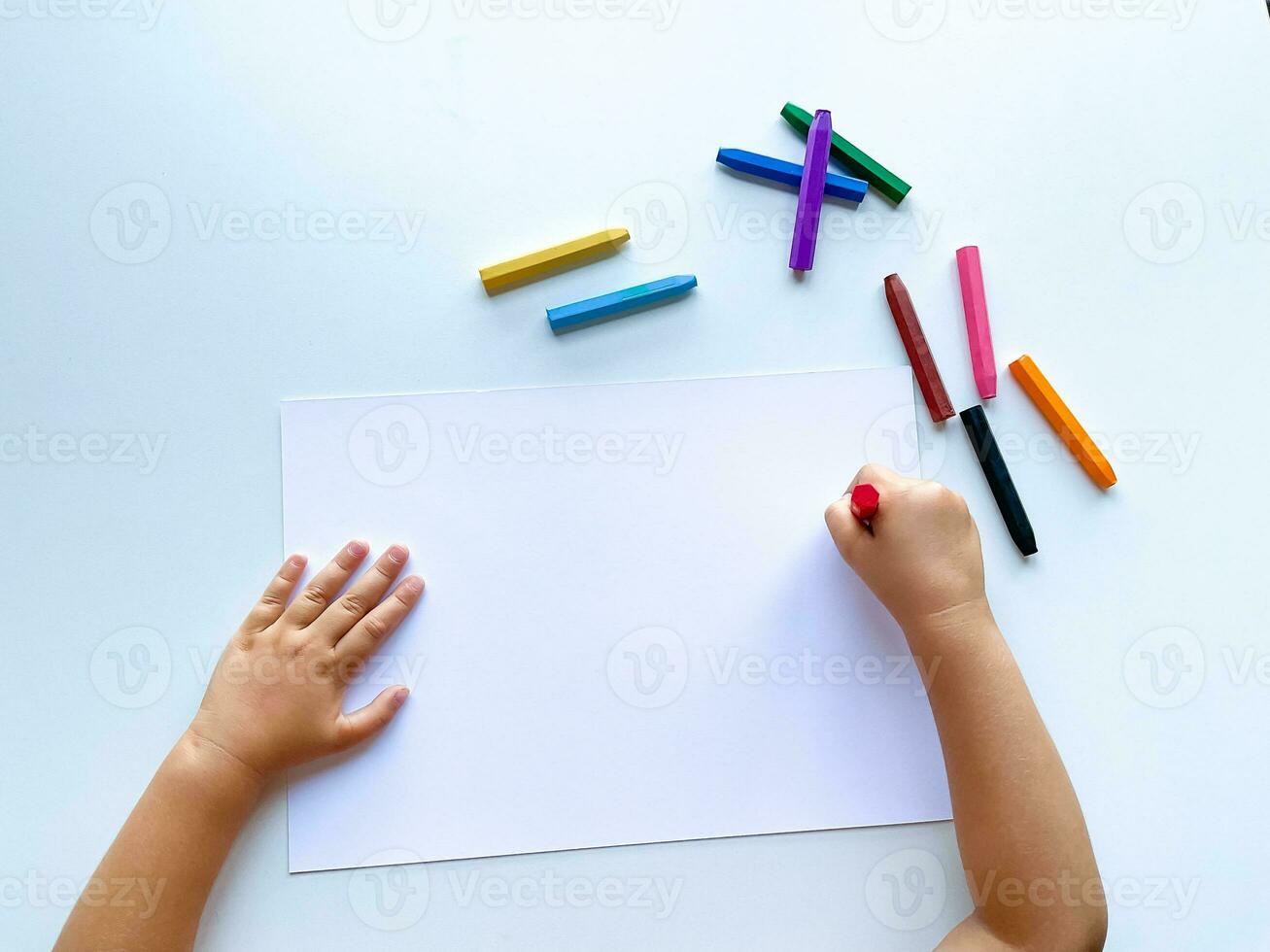 Childrens hands draw with colored wax crayons on a white sheet of paper. Top view of a blank sheet. photo