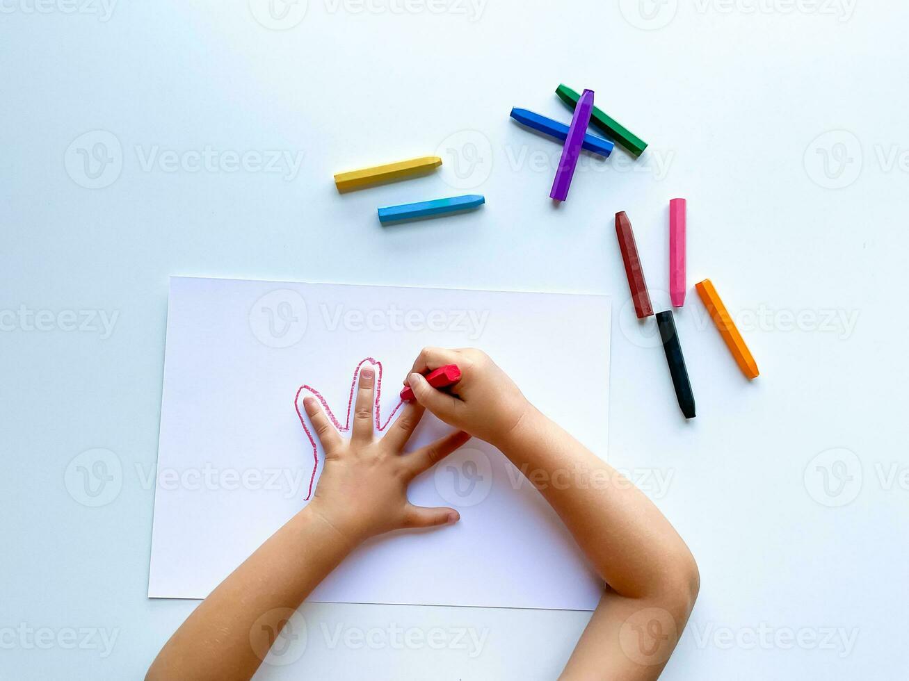 Childrens hands draw their hand with wax crayons on white paper, top view. photo