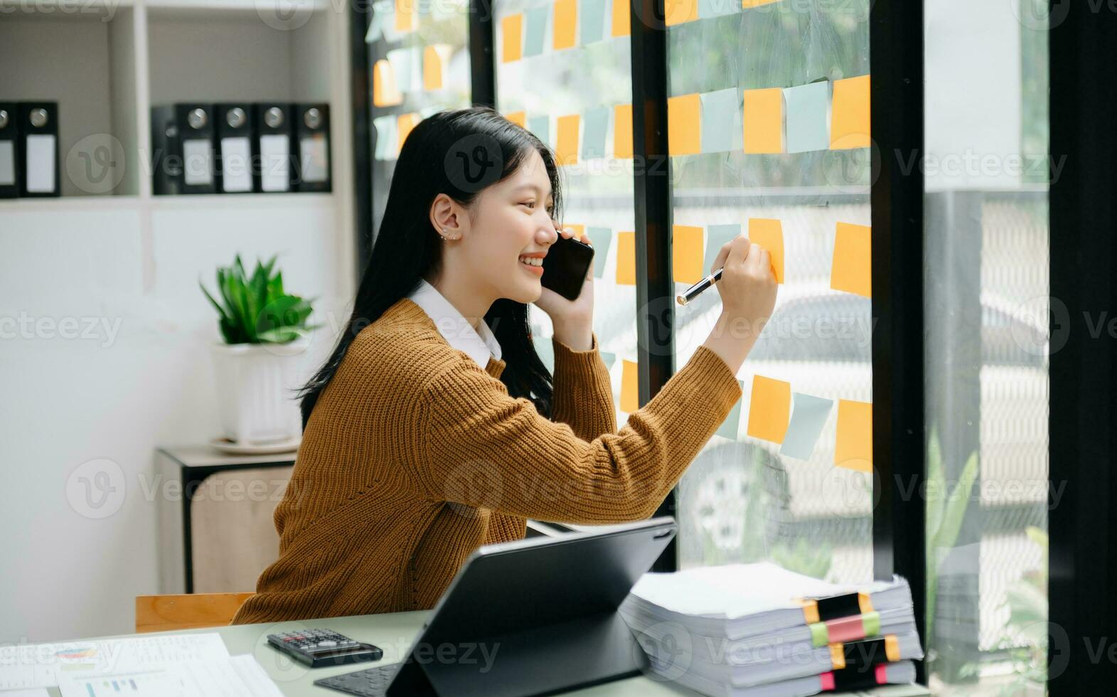 Asian businesswoman holding a pen taking note on note-paper attached to a mirror as a reminder laptop and tablet on the desk.in office photo