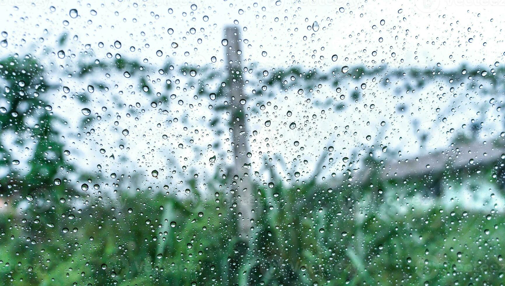 Rain droplets on surface of car glass with blurred green nature background and flowers through window glass of the car covered by raindrops. Freshness after rain. Wet windscreen shot from inside car. photo