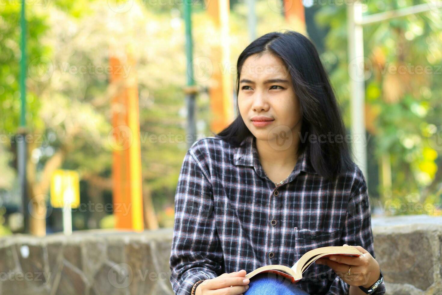 happy young asian college student read a book in the park looking away photo