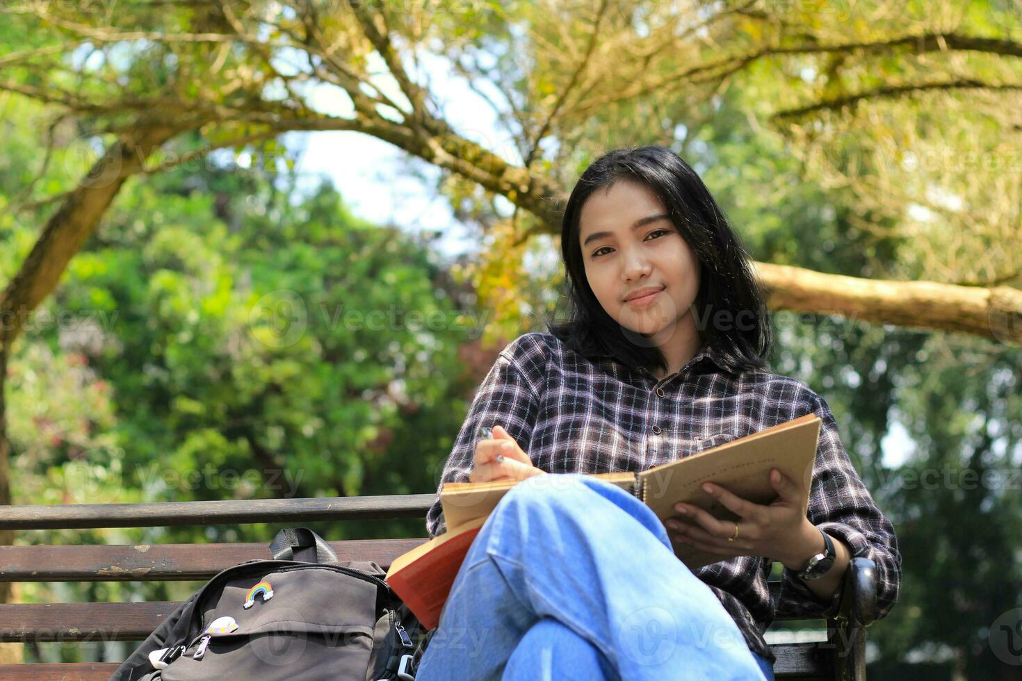sonriente atractivo asiático joven mujer escritura a hacer lista y organizando su trabajo en cuaderno en ciudad parque foto