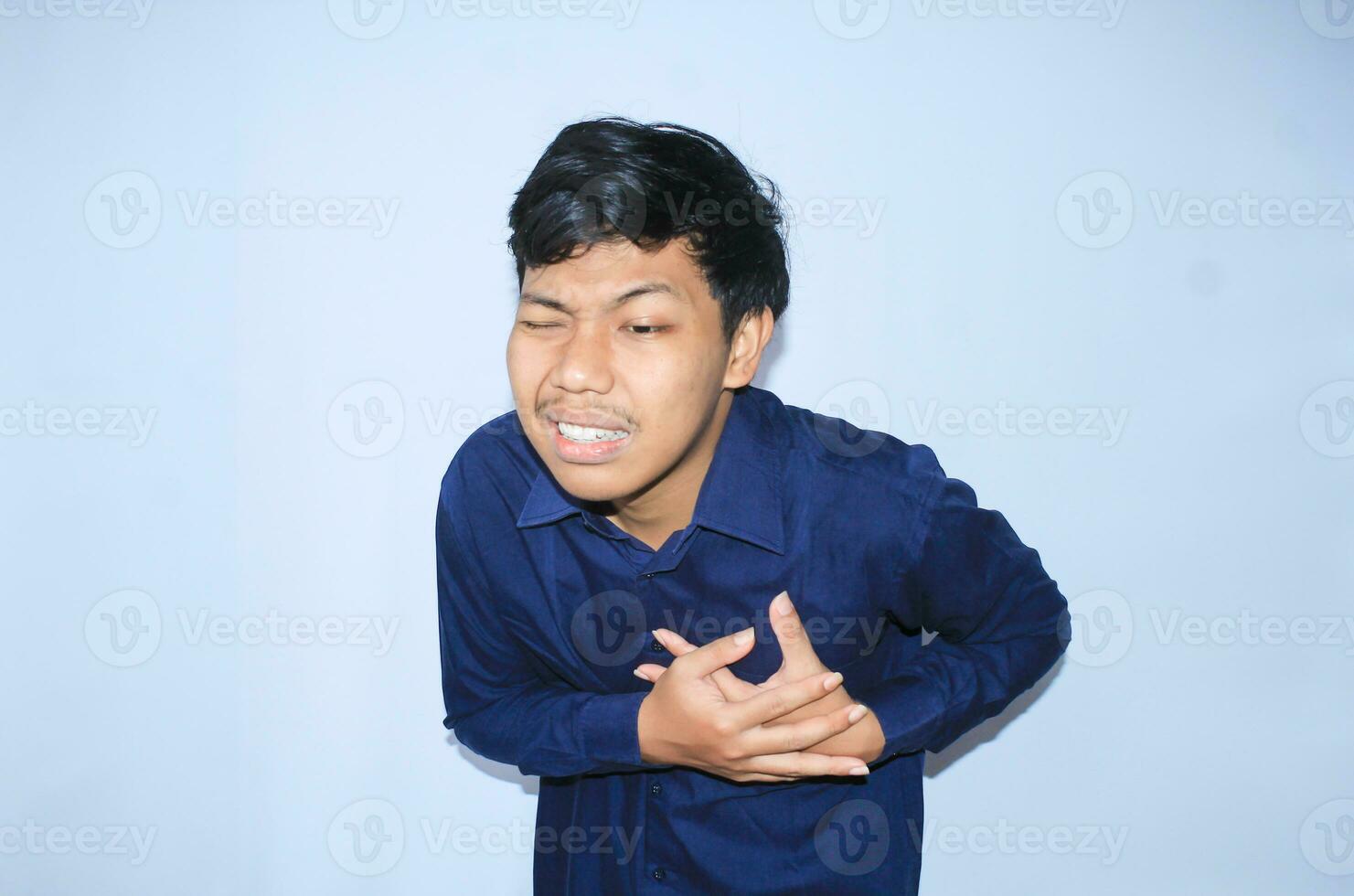 young asian man in pain of heart attack is holding his chest with two hands and closed one eye wearing navy shirt, isolated white photo