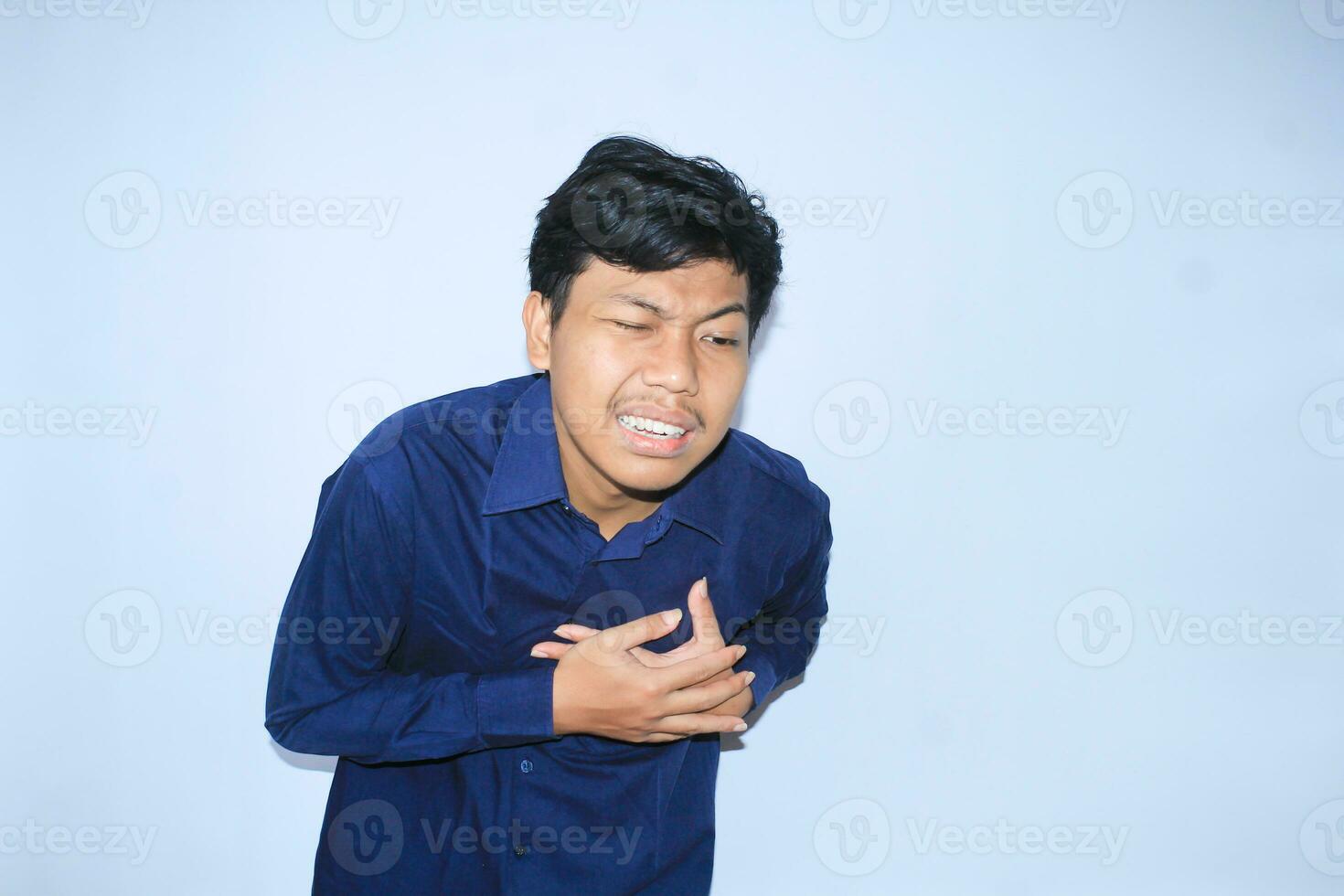 young asian man in pain of heart attack is holding his chest with two hands and closed one eye wearing navy shirt, isolated white photo