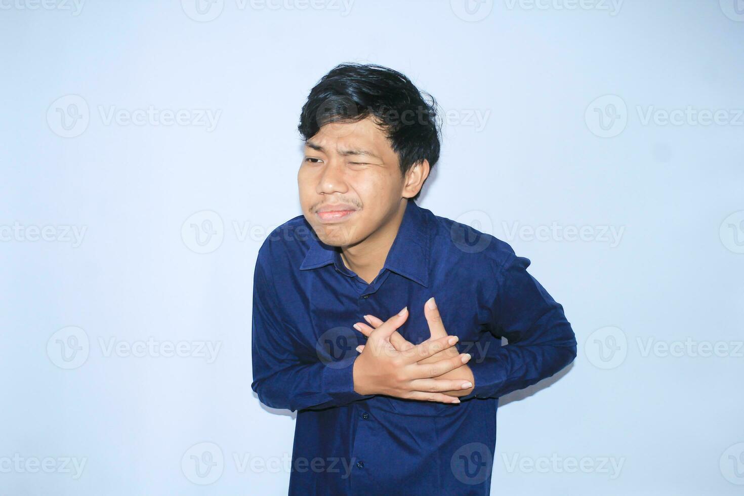 young asian man in pain of heart attack is holding his chest with two hands and closed one eye wearing navy shirt, isolated white photo