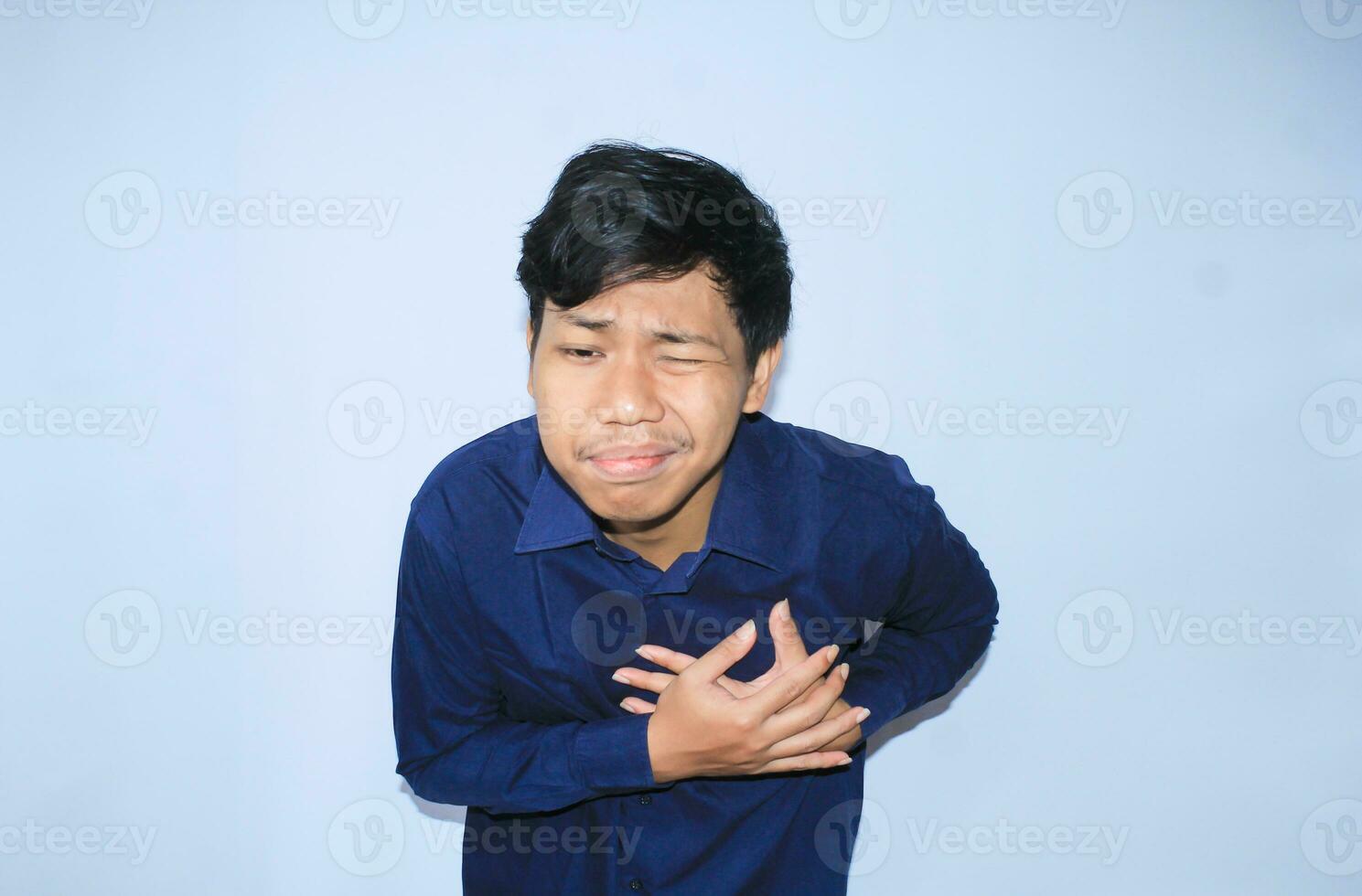 young asian man in pain of heart attack is holding his chest with two hands and closed one eye wearing navy shirt, isolated white photo