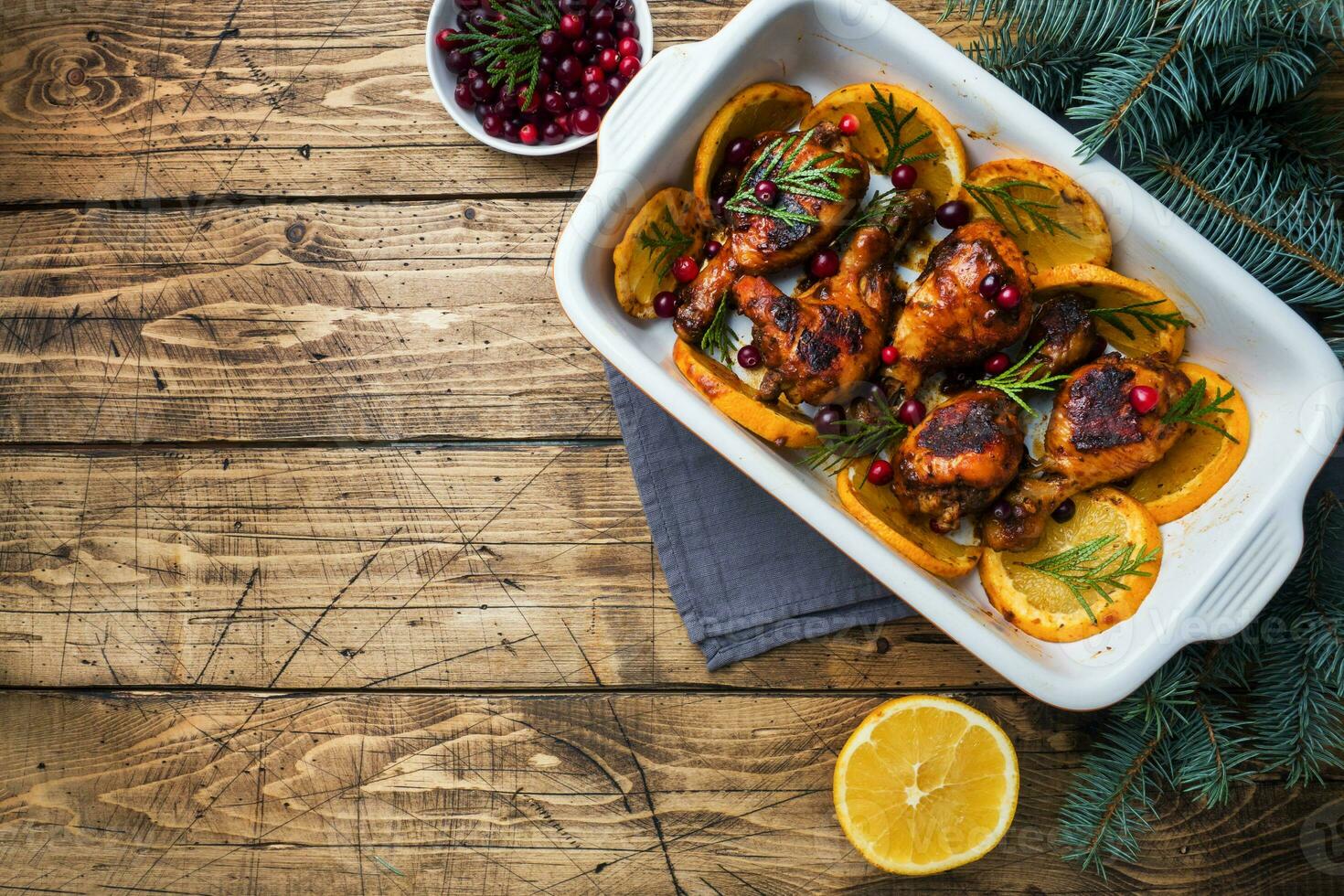 Baked chicken drumstick with oranges and cranberries in a baking sheet on a wooden background. Christmas food Table with decorations. Copy space. photo