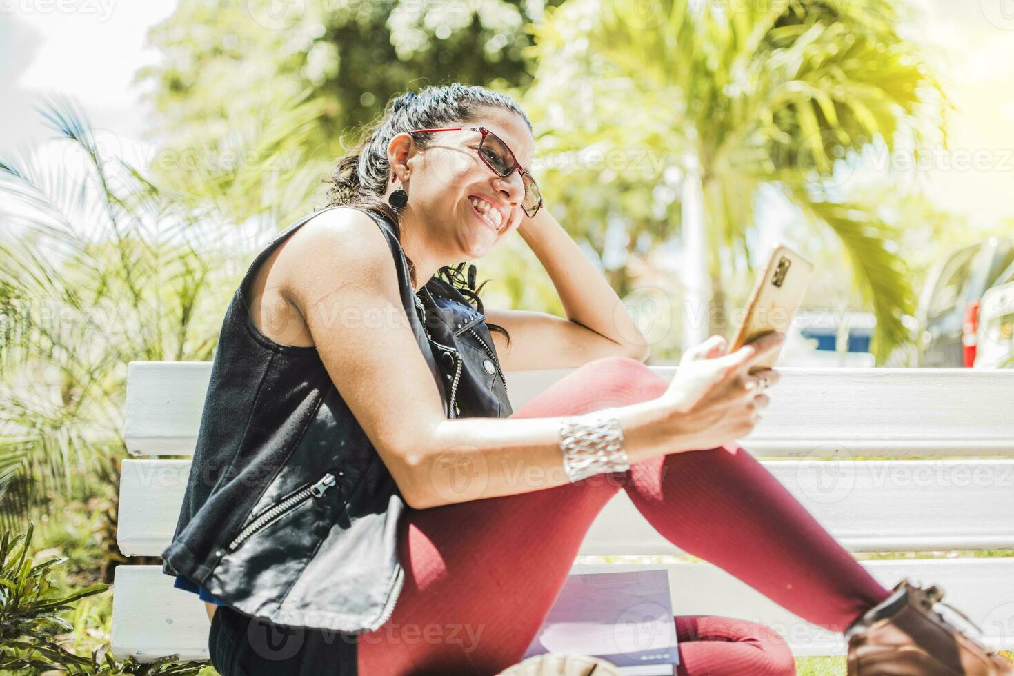 Girl sitting on a bench checking her cell phone, Happy woman sitting in a park texting on her cell phone, Happy woman sitting on a bench sending a text message photo