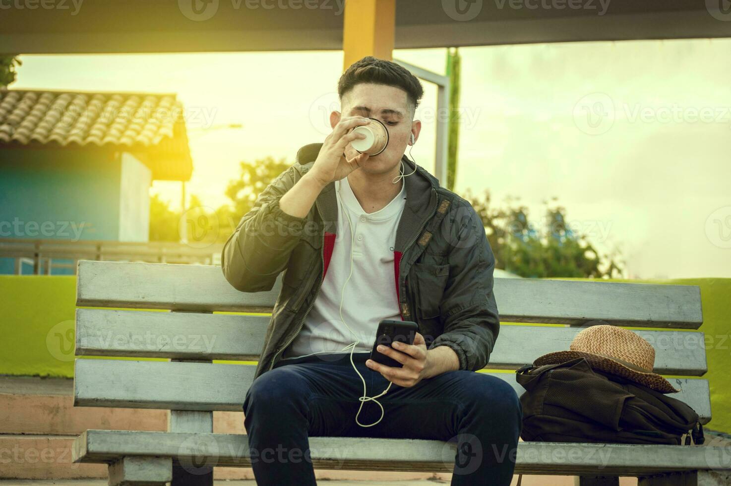 relajado atractivo chico escuchando a música en un banco, chico sentado en un banco escuchando a música y Bebiendo café foto