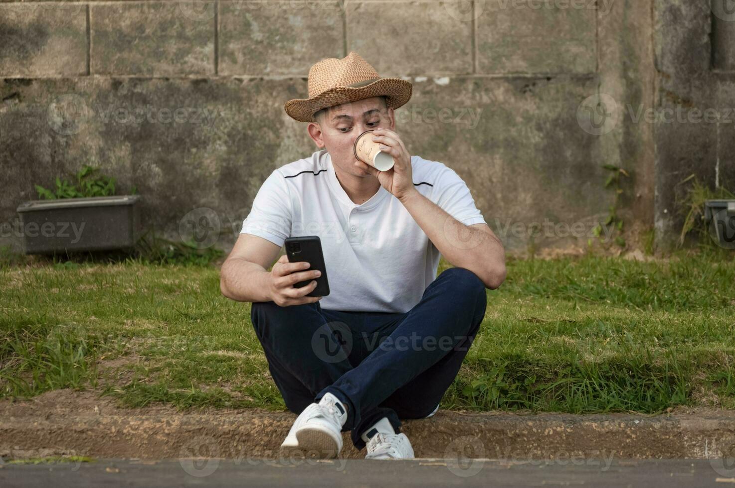 joven hombre sentado en acera comprobación teléfono, hombre Bebiendo café mientras comprobación su teléfono foto