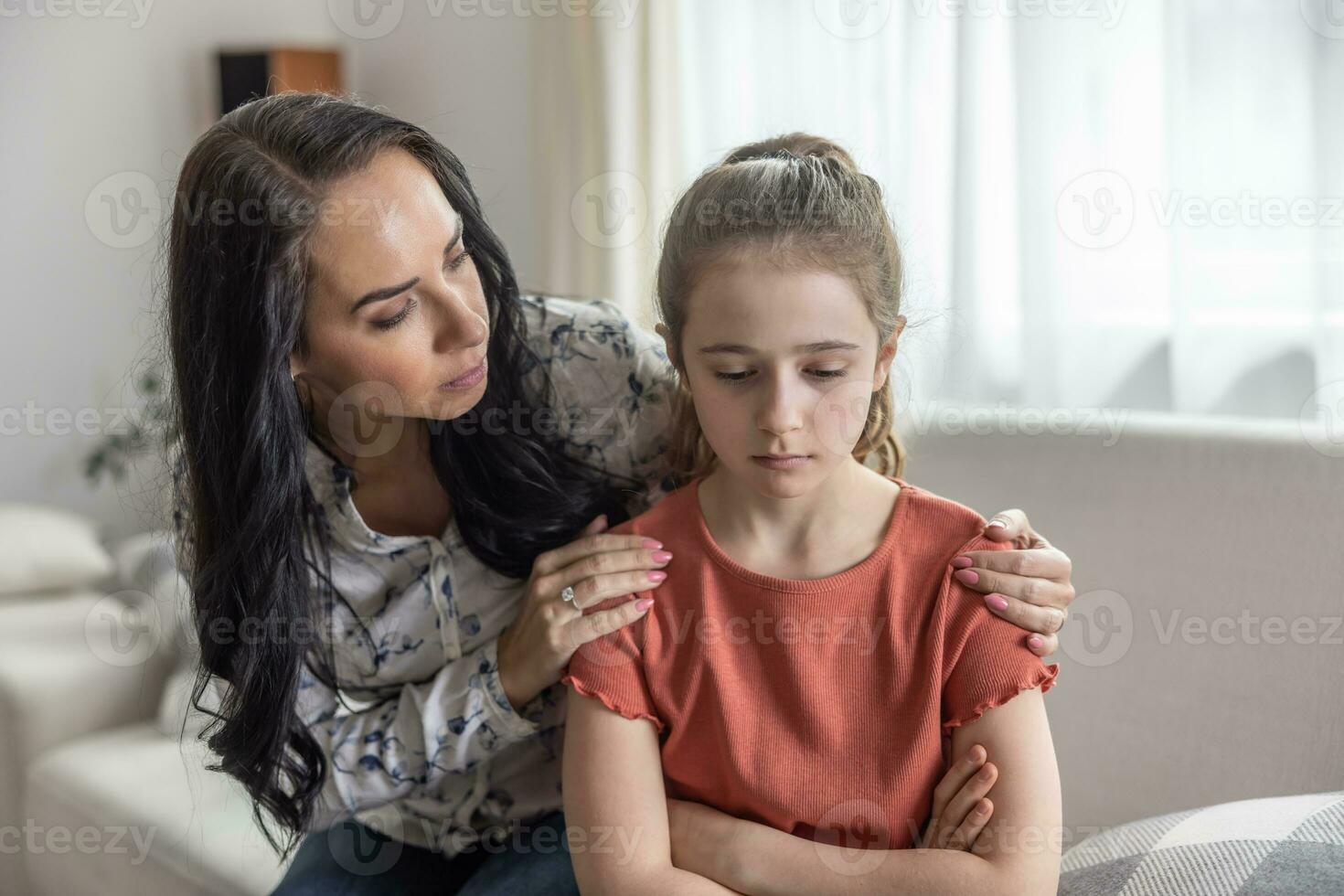 Mother comforts upset daughter that sits with arms crossed on a couch. photo