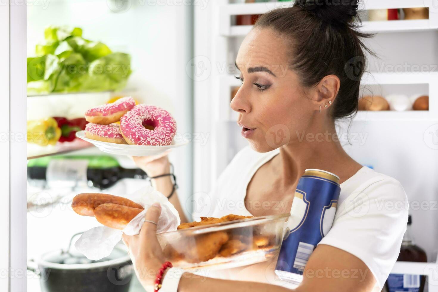 On Friday evening, a very hungry woman in pajamas chooses snacks, snitzels, donuts and a can of beer from the refrigerator photo