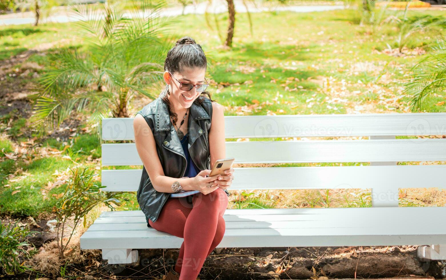 Girl sitting on a bench checking her cell phone, Happy woman sitting in a park texting on her cell phone, Woman sitting on a bench sending a text message photo