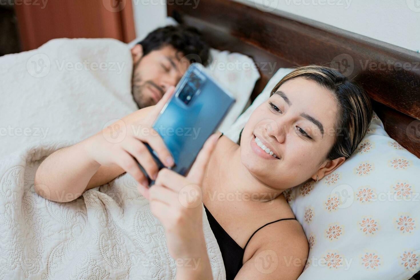 Unfaithful wife with phone while her husband sleeps. Unfaithful woman with phone while the man sleeps. Unfaithful girlfriend with phone while boyfriend sleeps photo