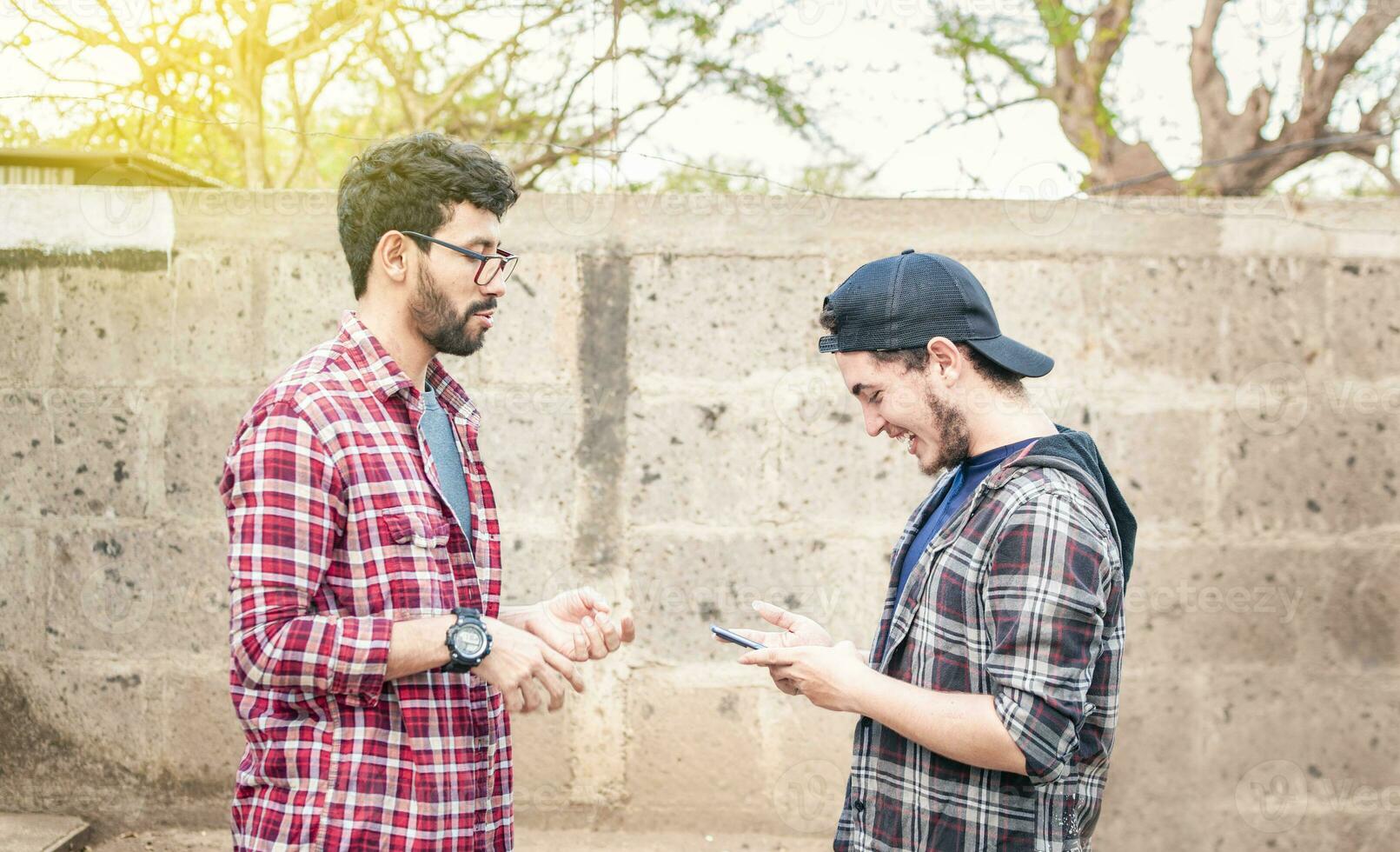 lado ver de dos joven amigos chateando al aire libre, dos amigos chateando mientras sonriente y utilizando Teléfono móvil. ver de dos adolescentes hablando cara a cara mientras utilizando el célula teléfono foto