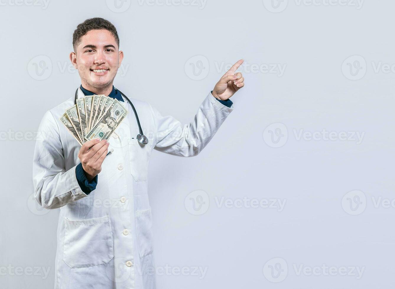 Happy latin doctor holding money and pointing to side. Handsome smiling doctor showing money and pointing side photo